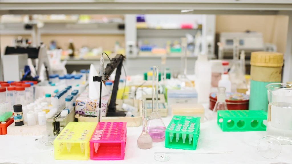 A researcher's gloved hand holds a small blood sample vial on a laboratory bench lit by warm window light.